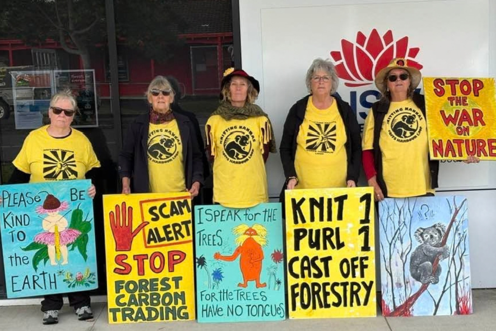 Five women wearing yellow Coffs Harbour Knitting Nannas t-shirts stand in a line outdoors holding colorful handwritten protest signs. The signs display messages including "PLEASE BE KIND TO THE EARTH", "SCAM ALERT: STOP FOREST CARBON TRADING", "I SPEAK FOR THE TREES FOR THE TREES HAVE NO TONGUES", "KNIT 1 PURL 1 CAST OFF FORESTRY", and "STOP THE WAR ON NATURE."